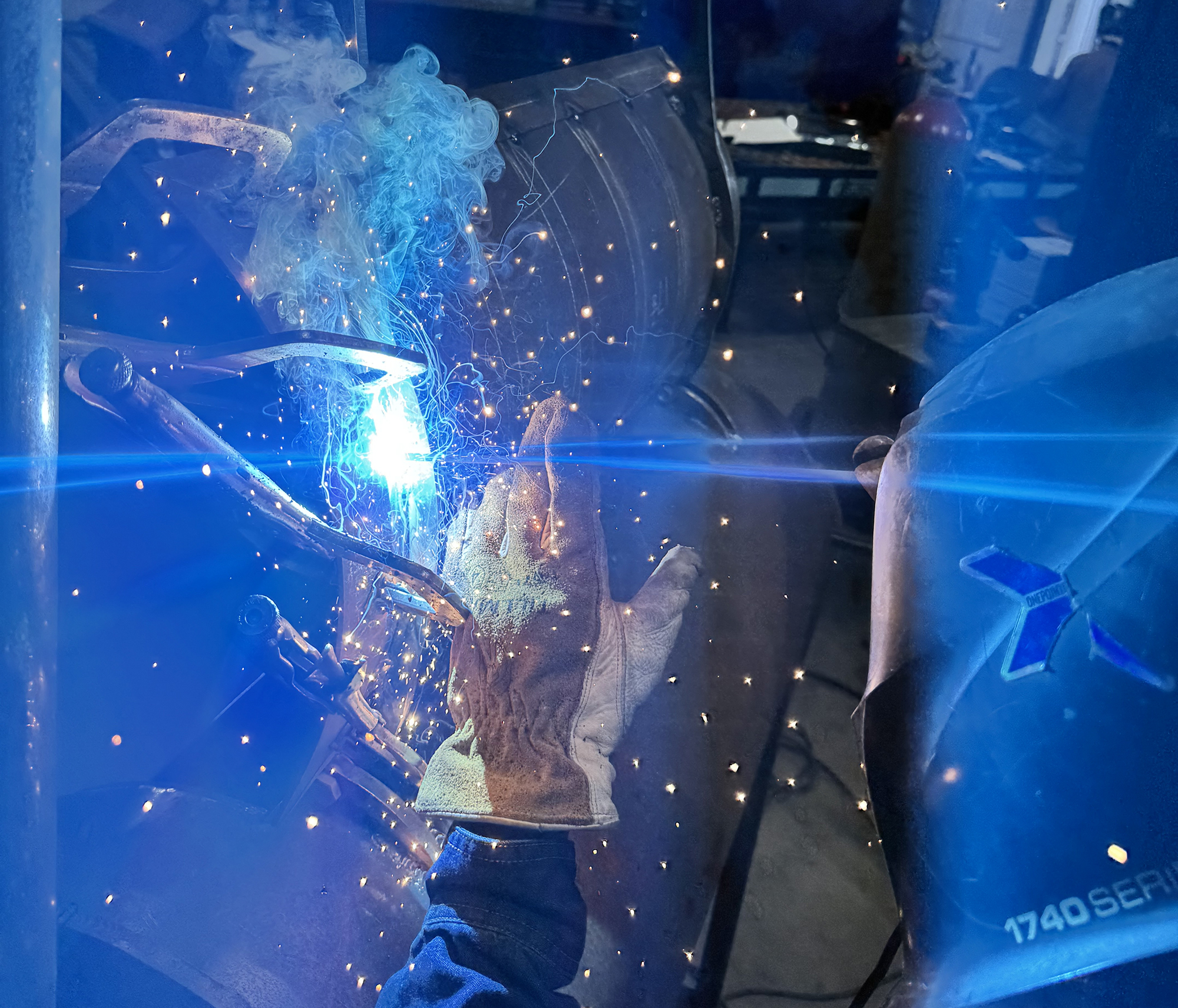 A student wearing protective welding gear and a helmet using a torch in a welding workshop, with sparks flying.